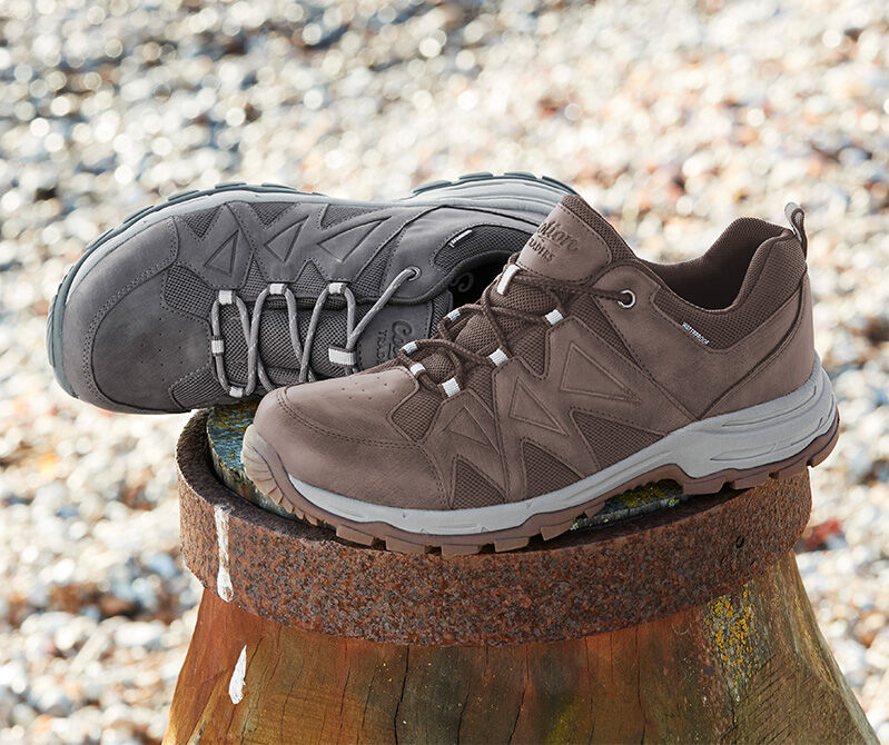 A still shot photograph of a pair of the Cotton Traders Hydroguard® Panel Detail Walking Shoes on a stoney beach