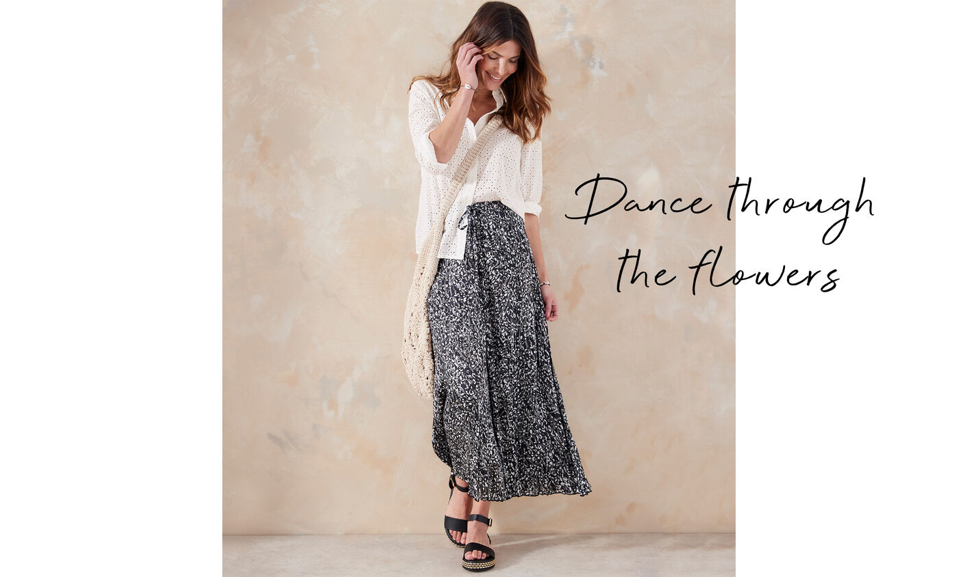 A woman is fixing her hair and looking down toward the ground. She wears a Cotton Traders printed maxi skirt with a broderie anglaise style blouse. Draped over her shoulder is a crossbody crochet bag. The text on this image reads 