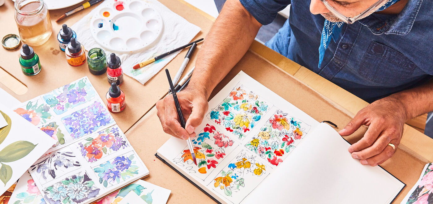 A man paints a floral design in a book while he’s surrounded by painting supplies, including ink, paintbrushes and a mixing palette