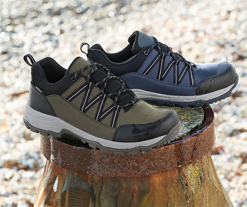 A still shot photograph of a pair of the Cotton Traders Hydroguard® Tape Detail Walking Shoes on a stoney beach