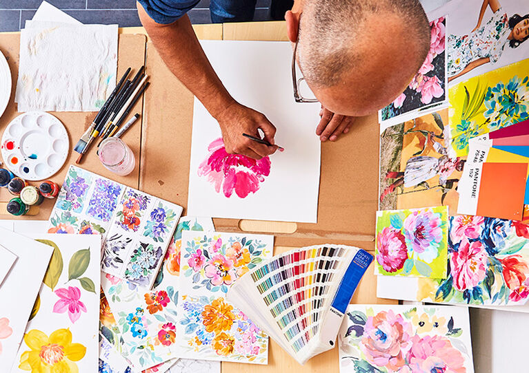 An overhead shot of the man painting a pink flower. He is surrounded by other floral paintings as well as a mixing palette, paintbrushes, paints and inks
