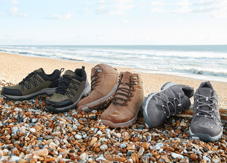 A collection of the Cotton Traders Hydroguard® Waterproof Walking Shoes and Boots set in a row on a stoney beach