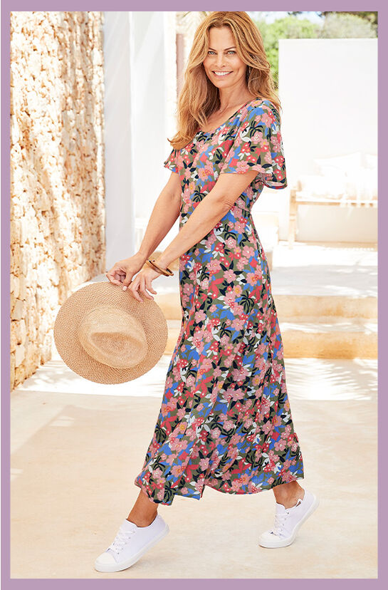 A woman wears a floral maxi dress with flutter sleeves. She pairs these with a pair of white trainers and has a sun hat in her hand.