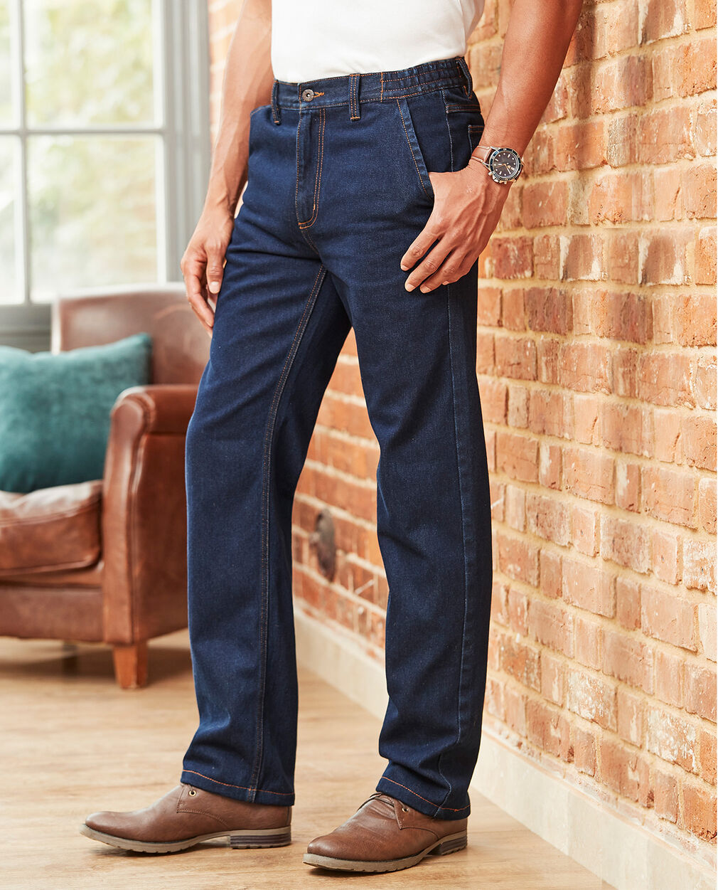 Man wearing a pair of dark blue wash denim jeans with a white T-shirt tucked in and brown shoes. The background is of a brick wall