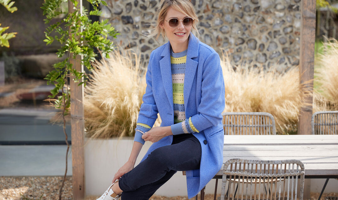 A woman is outside sitting on the corner of a table. She is wearing a pastel blue, pink, yellow and green knitted jumper with dark wash jeans and a light blue coat on top. She is also wearing oversized sunglasses