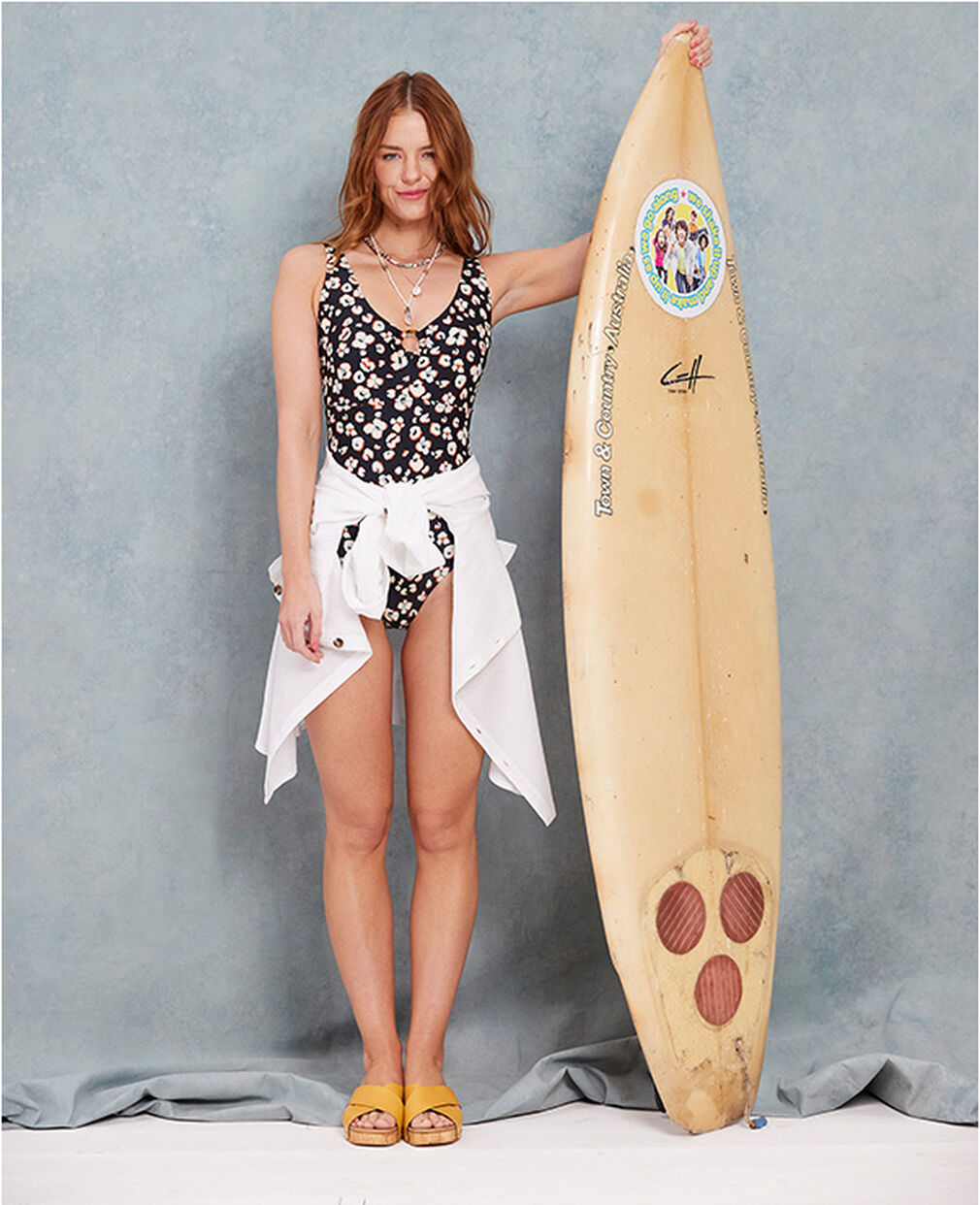 A woman stands next to a surfboard. She wears a black floral swimming costume with a white shirt tied around her waist. She also wears a pair of yellow sandals.