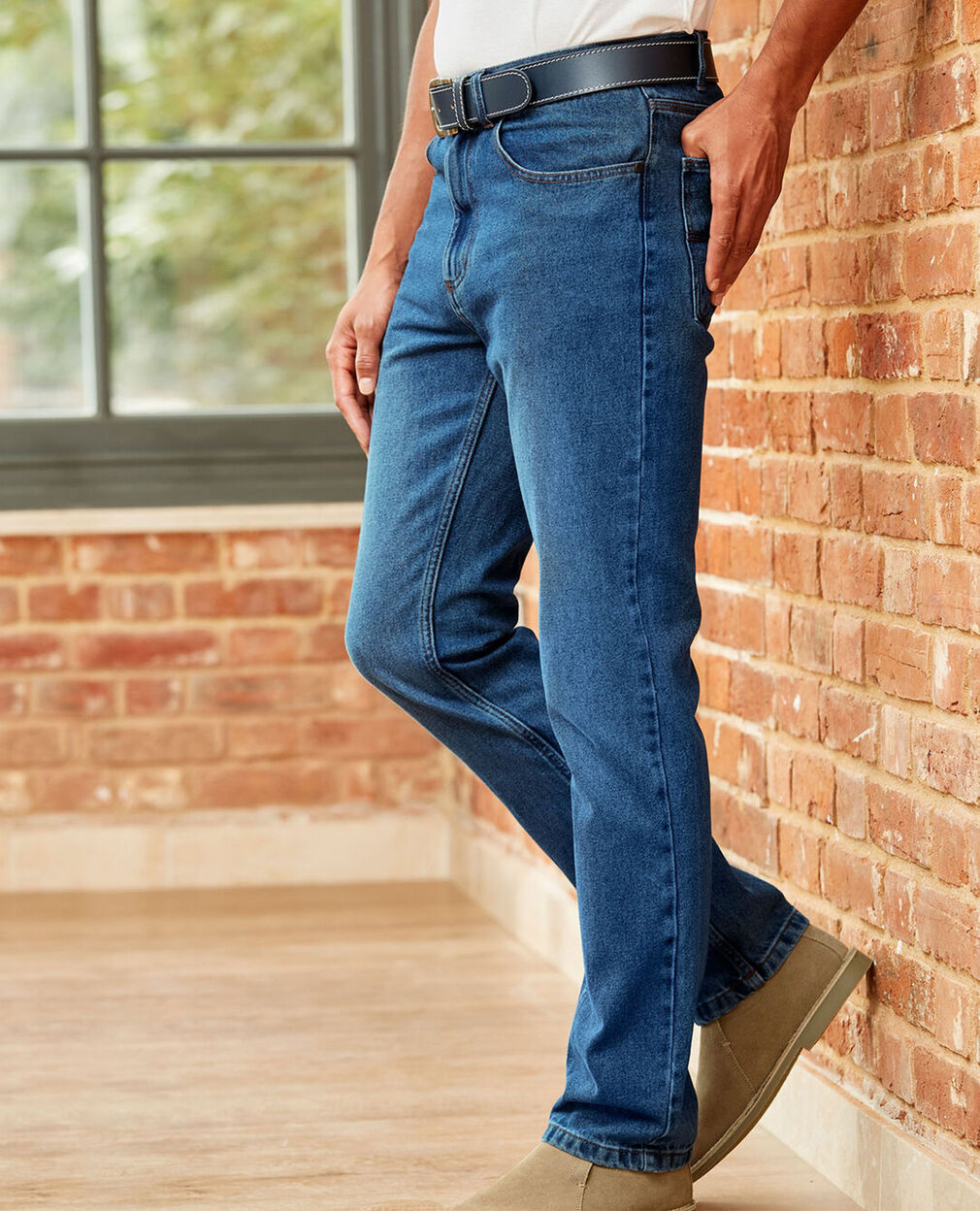 Man wearing a pair of mid-wash blue jeans with a black belt and a white T-shirt tucked in. He is also wearing a pair of brown boots and the backdrop is of a brick wall