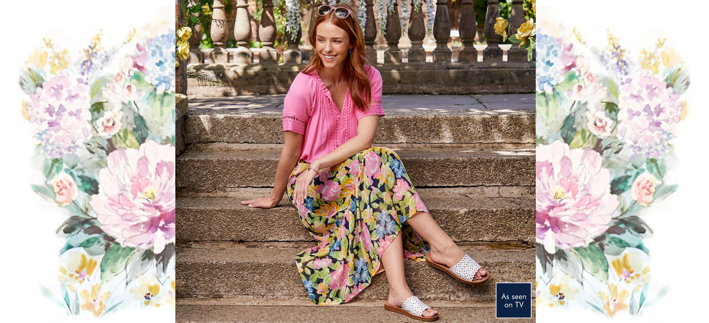 A woman is sitting on steps wearing a pink blouse with a floral midi skirt and white sandals